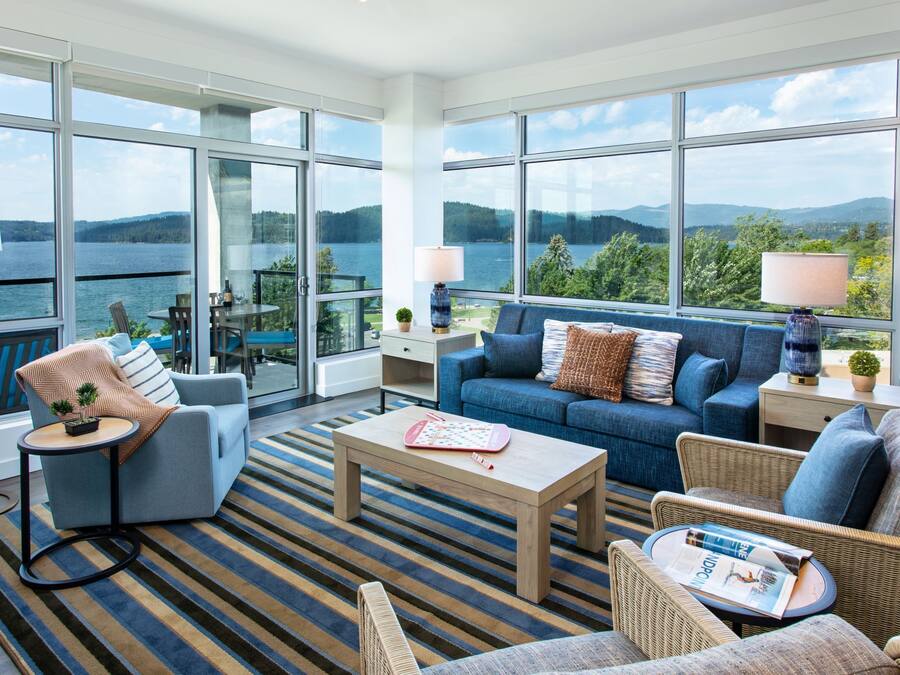 A spacious living room with large windows offering a view of a lake and mountains. There is a blue sofa, a coffee table, and wicker chairs.