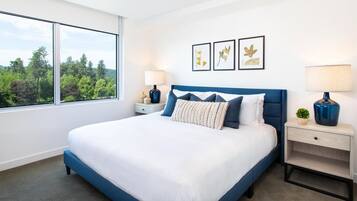 Une chambre moderne avec un grand lit, deux tables de chevet, une petite table et une vue sur les arbres par une grande fenêtre.