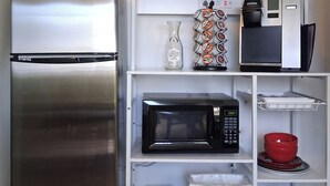 A compact kitchen with a stainless steel refrigerator, a microwave, a coffee maker, and a kettle on the shelves.
