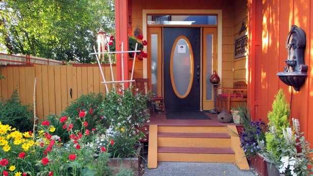 A wooden house with a red door, a small porch, and a garden with various flowers.