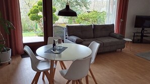 A living room with a round table, white chairs, a grey sofa, a TV, and a view of greenery outside. - Ferienwohnung Schöne Aussicht (Weilrod)