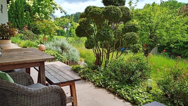An outdoor patio with a wooden table and wicker chairs, surrounded by lush greenery and trees. - Ferienwohnung Schöne Aussicht (Weilrod)