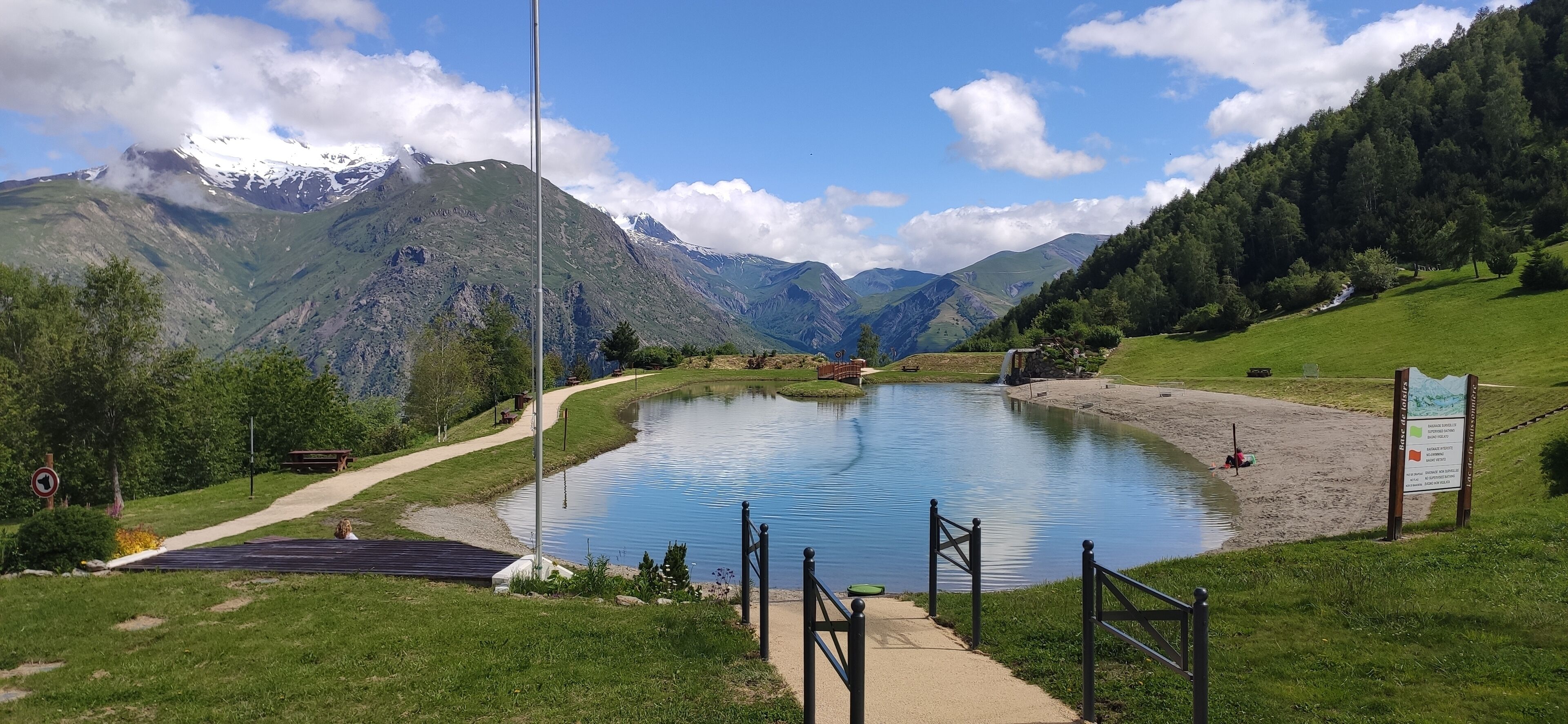 Een meer met een houten terras en een picknicktafel, omringd door bergen en een heldere lucht.