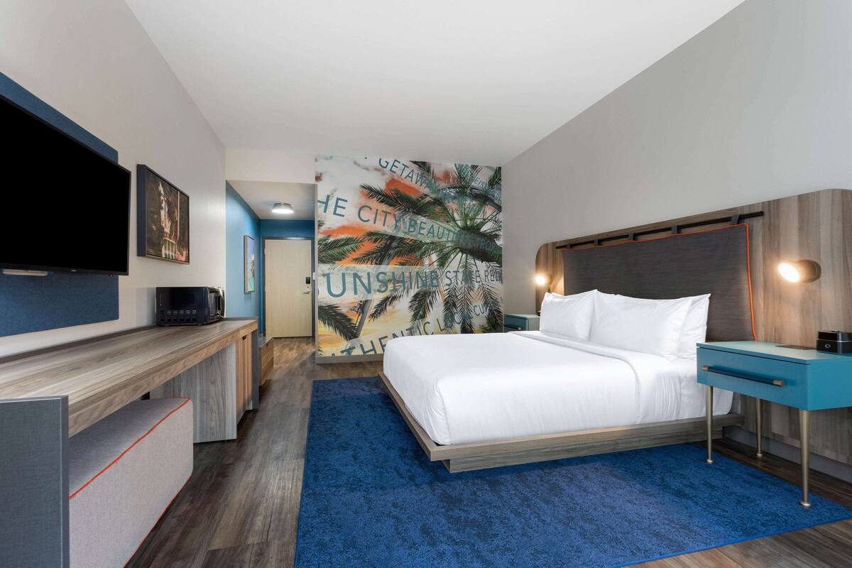 Una habitación de hotel moderna con una cama grande, un televisor de pantalla plana, un escritorio y una pared con un mural tropical.