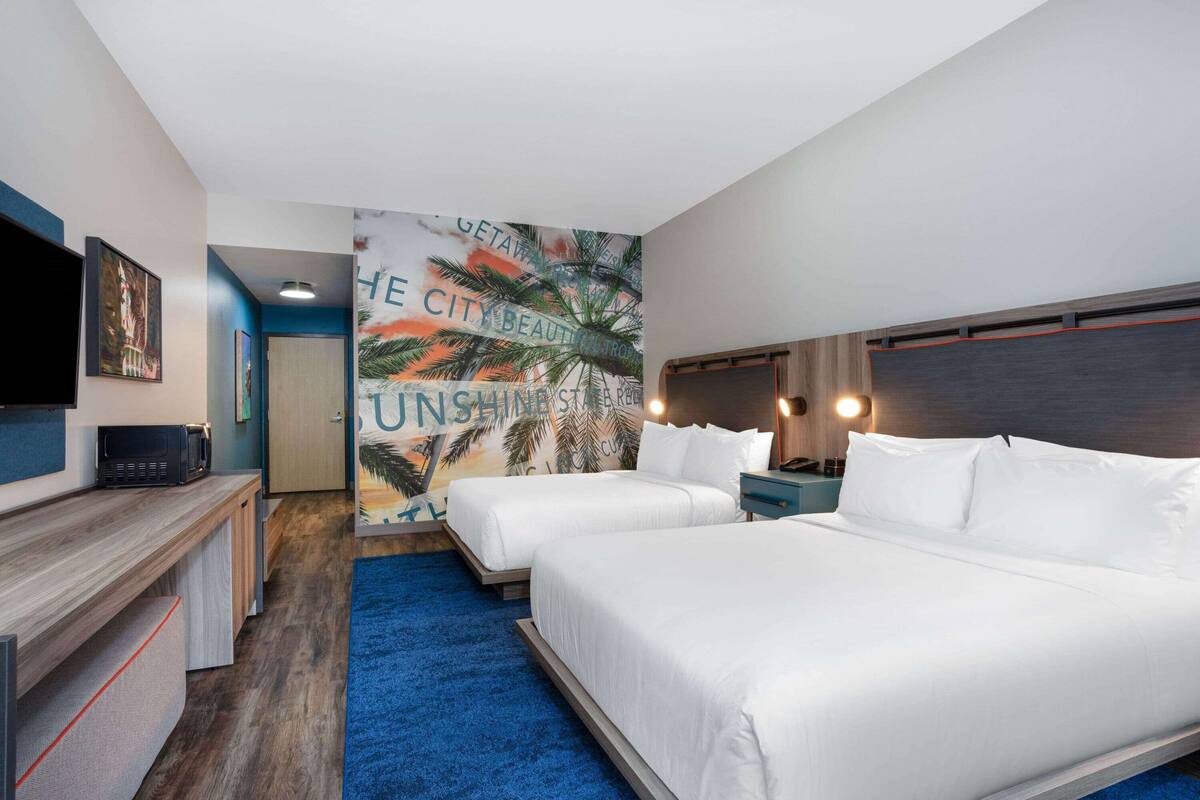 Habitación de hotel con dos camas, televisión, escritorio y un mural con palmeras y texto.