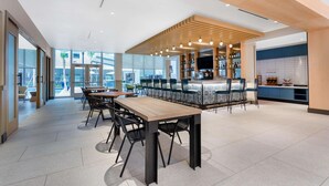 A modern dining area with a long wooden table and black chairs, a bar area with a patterned backsplash, and a view of the outside through large windows.