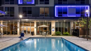 A modern hotel with a swimming pool, outdoor seating, and a well-lit facade.