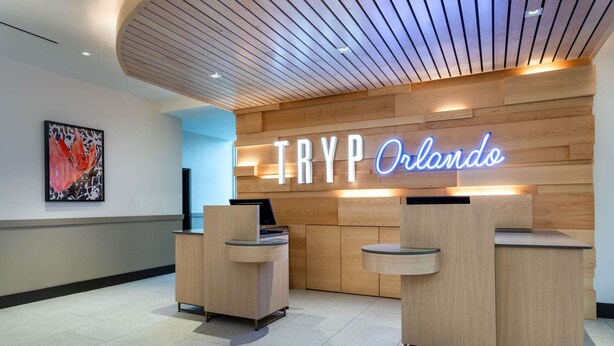 A reception desk area with a neon sign reading "TRYP Orlando" and a modern wooden ceiling.
