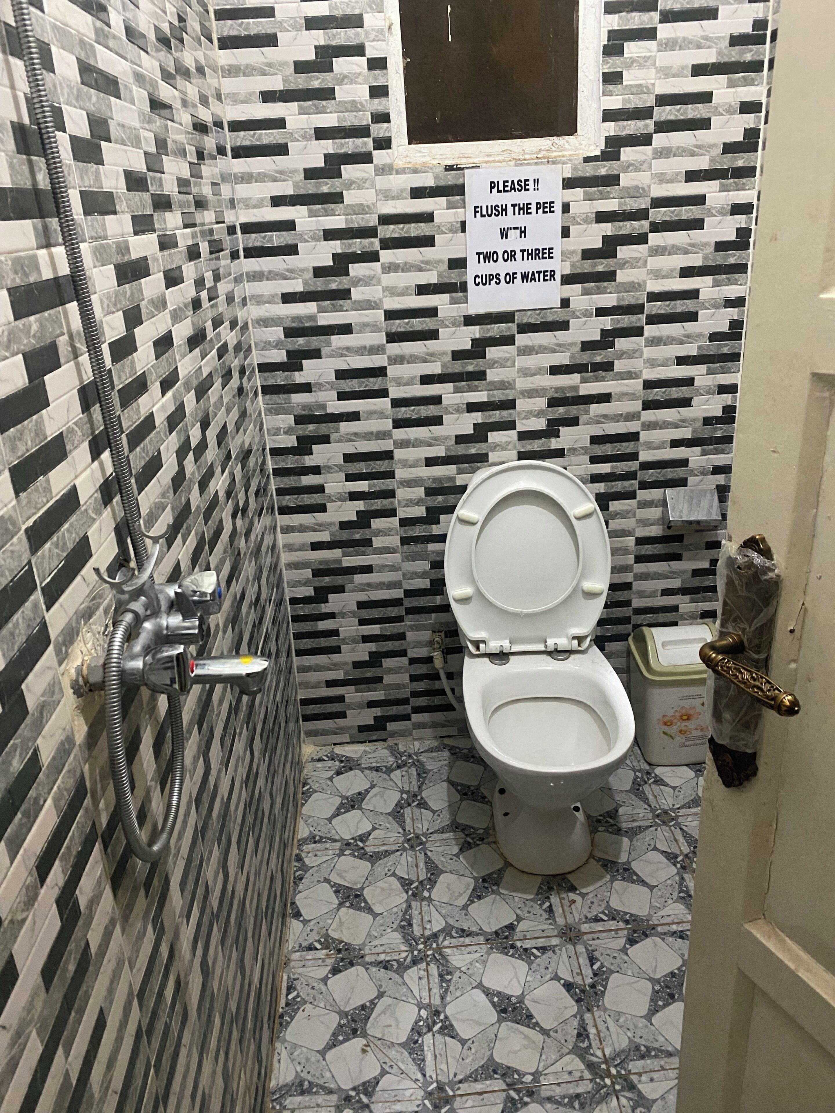 Un baño con piso de baldosas, un inodoro, una ducha con manguera y un cartel que indica tirar agua.