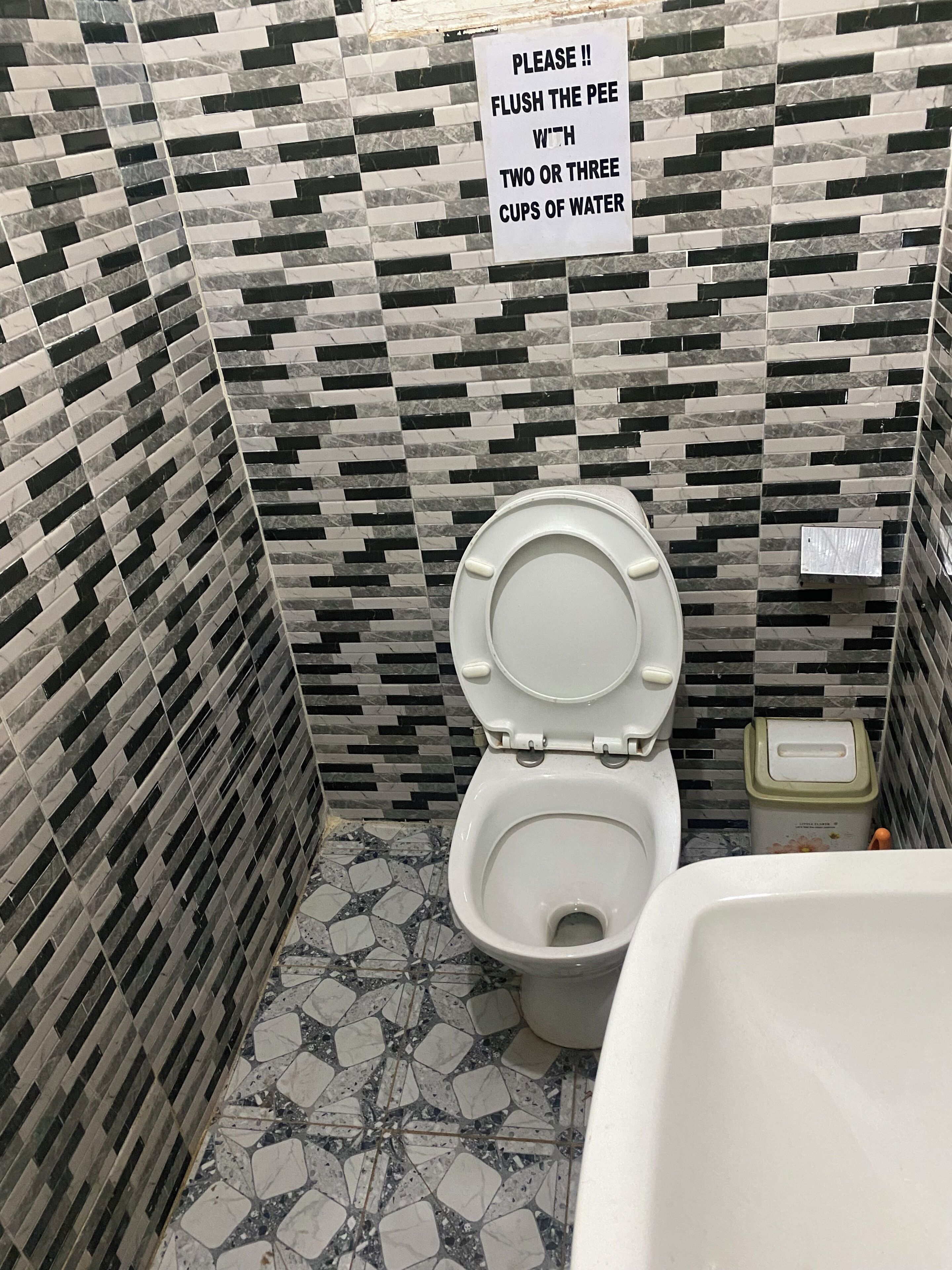 A bathroom with a toilet, a trash bin, and a sign instructing to flush with two or three cups of water.