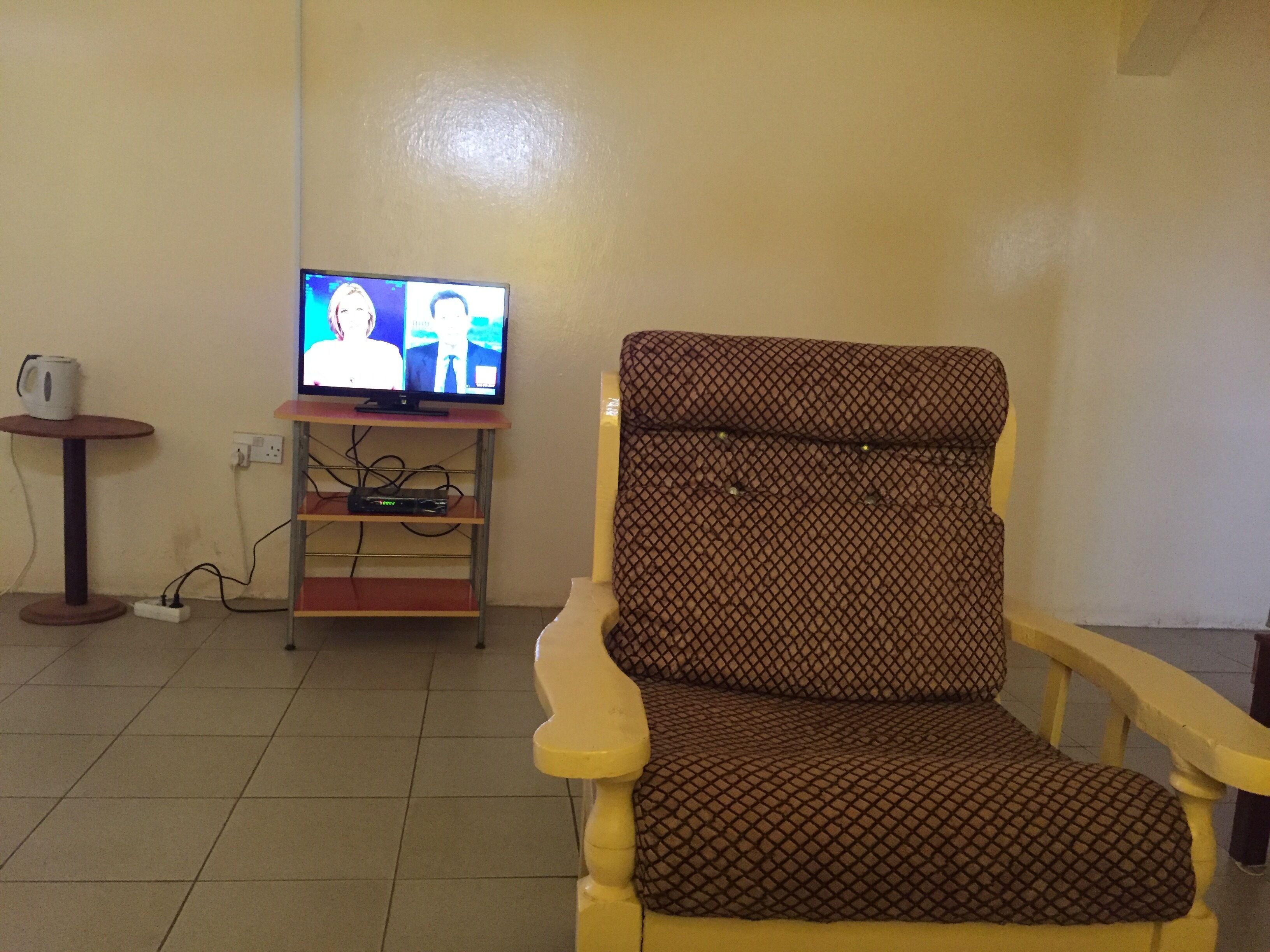 Sala de estar com televisão em um suporte, poltrona estampada, uma mesinha com uma caneca e uma tomada.