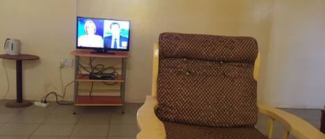 Sala de estar com televisão em um suporte, poltrona estampada, uma mesinha com uma caneca e uma tomada.