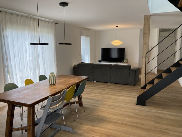 Uma sala de estar moderna com uma mesa de jantar de madeira, cadeiras coloridas, um sofá cinzento e uma TV em um suporte. Uma escada com corrimão de madeira leva a outro nível.