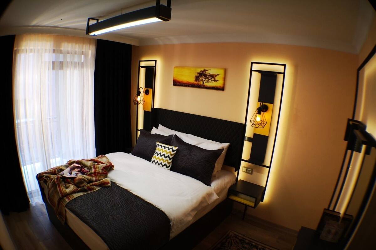 A modern hotel room with a large bed, a bedside table, a nightstand, a wall-mounted lamp, and a painting.
