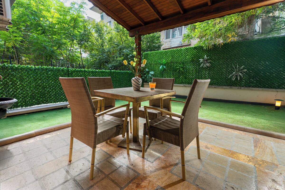 A covered patio with a table and chairs, a vase with flowers, and a view of a well-maintained garden.