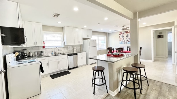 A modern kitchen with white cabinets, a stainless steel refrigerator, a microwave, and a coffee maker. There is a dining area with a red table and two bar stools. - BodhiTree Home - 3 Bedroom Bungalow (Houston)