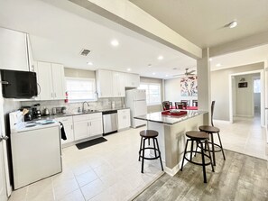 A modern kitchen with white cabinets, a stainless steel refrigerator, a microwave, and a coffee maker. There is a dining area with a red table and two bar stools. - BodhiTree Home - 3 Bedroom Bungalow (Houston)
