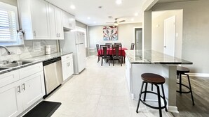 A modern kitchen with white cabinets, a refrigerator, a dining area with a red tablecloth, and a bar area with stools. - BodhiTree Home - 3 Bedroom Bungalow (Houston)