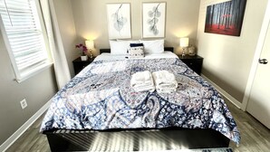 A neatly made bed with a patterned bedspread, two bedside tables with lamps, and a painting on the wall. - BodhiTree Home - 3 Bedroom Bungalow (Houston)