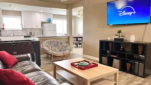 A living room with a leather sofa, a wooden coffee table, a wicker chair, a flat-screen TV displaying Disney+, and a kitchen in the background. - BodhiTree Home - 3 Bedroom Bungalow (Houston)