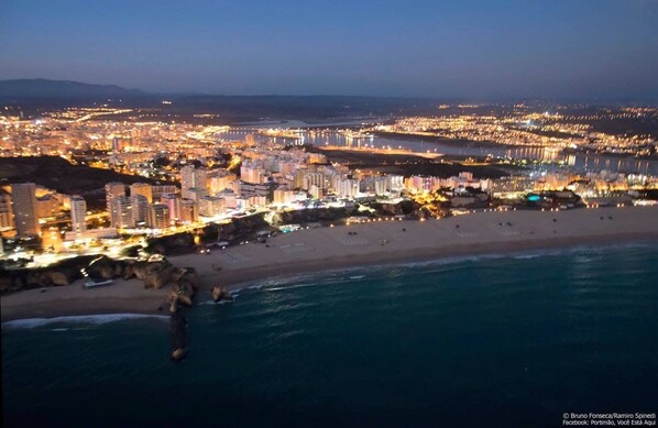A coastal cityscape at dusk with illuminated buildings, a beach, and a body of water.