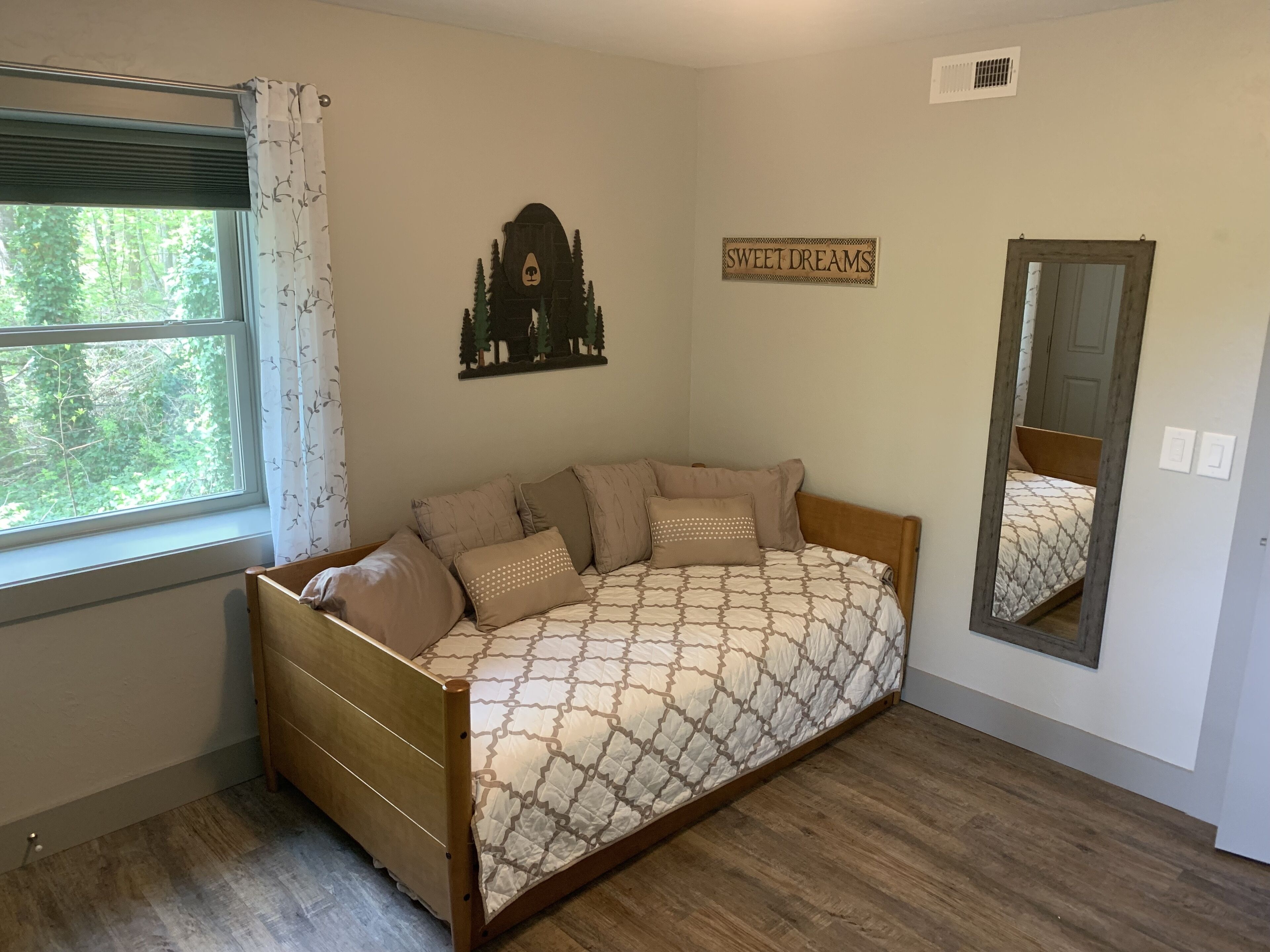 A bedroom with a wooden bed, a window with curtains, a wall-mounted mirror, and a 'SWEET DREAMS' sign.