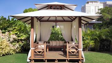 Un gazebo in legno con tettoia, sedute imbottite e vista su vegetazione rigogliosa e un edificio sullo sfondo.
