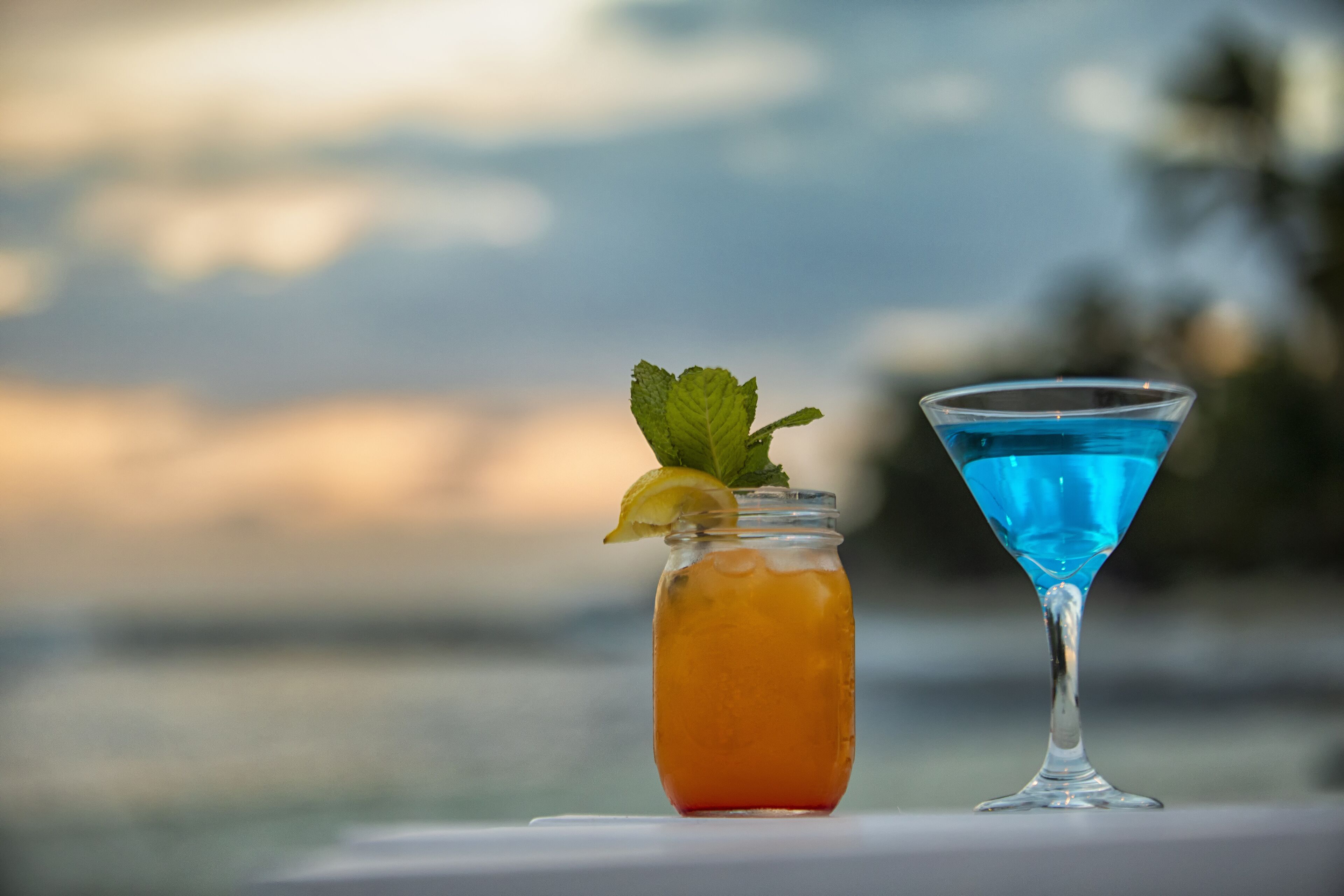 Dos bebidas, una en un tarro y otra en una copa de martini, con un fondo desenfocado al aire libre.
