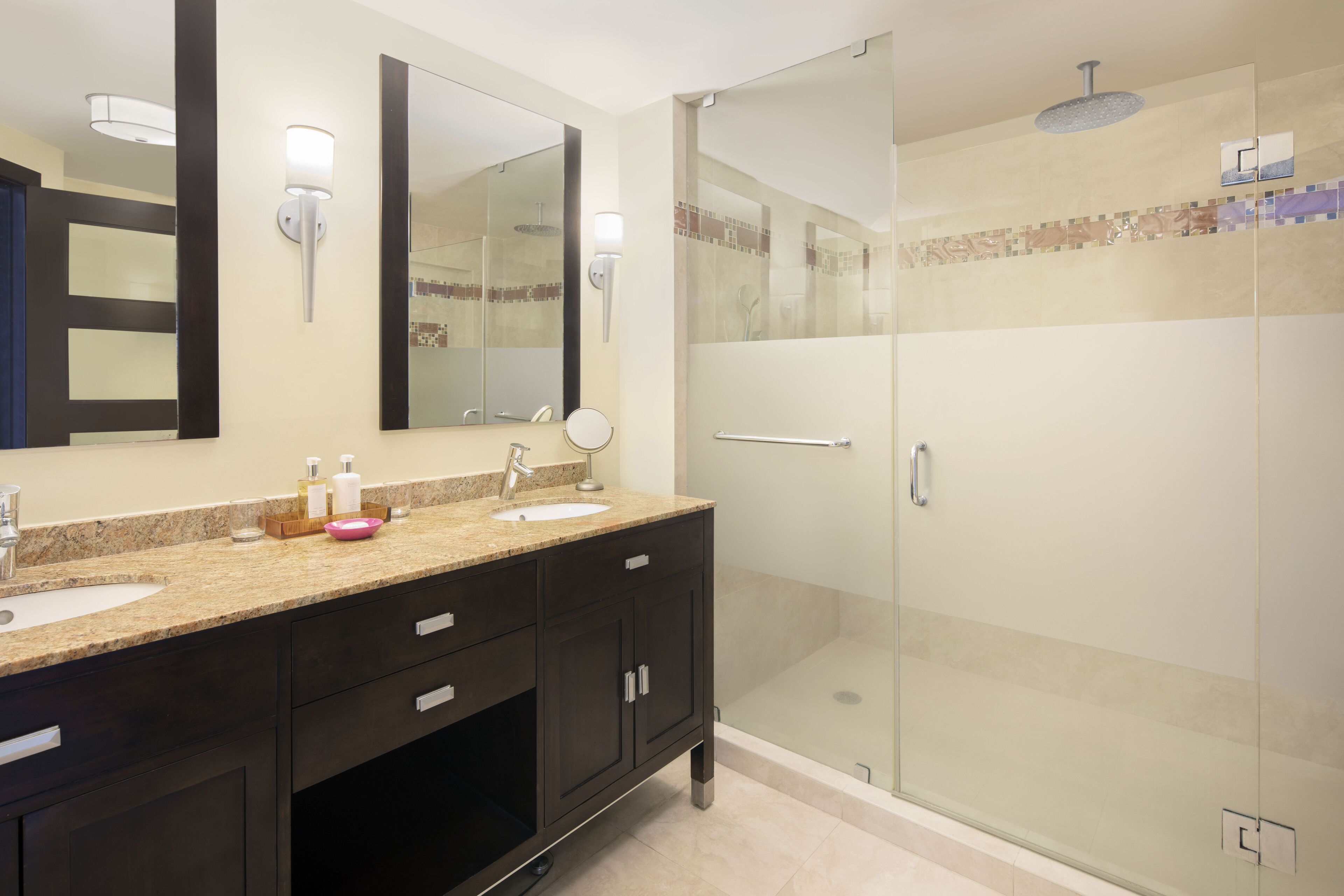 Un baño moderno con un espejo grande, dos lavamanos, muebles de color oscuro y una zona de ducha con mampara de vidrio.