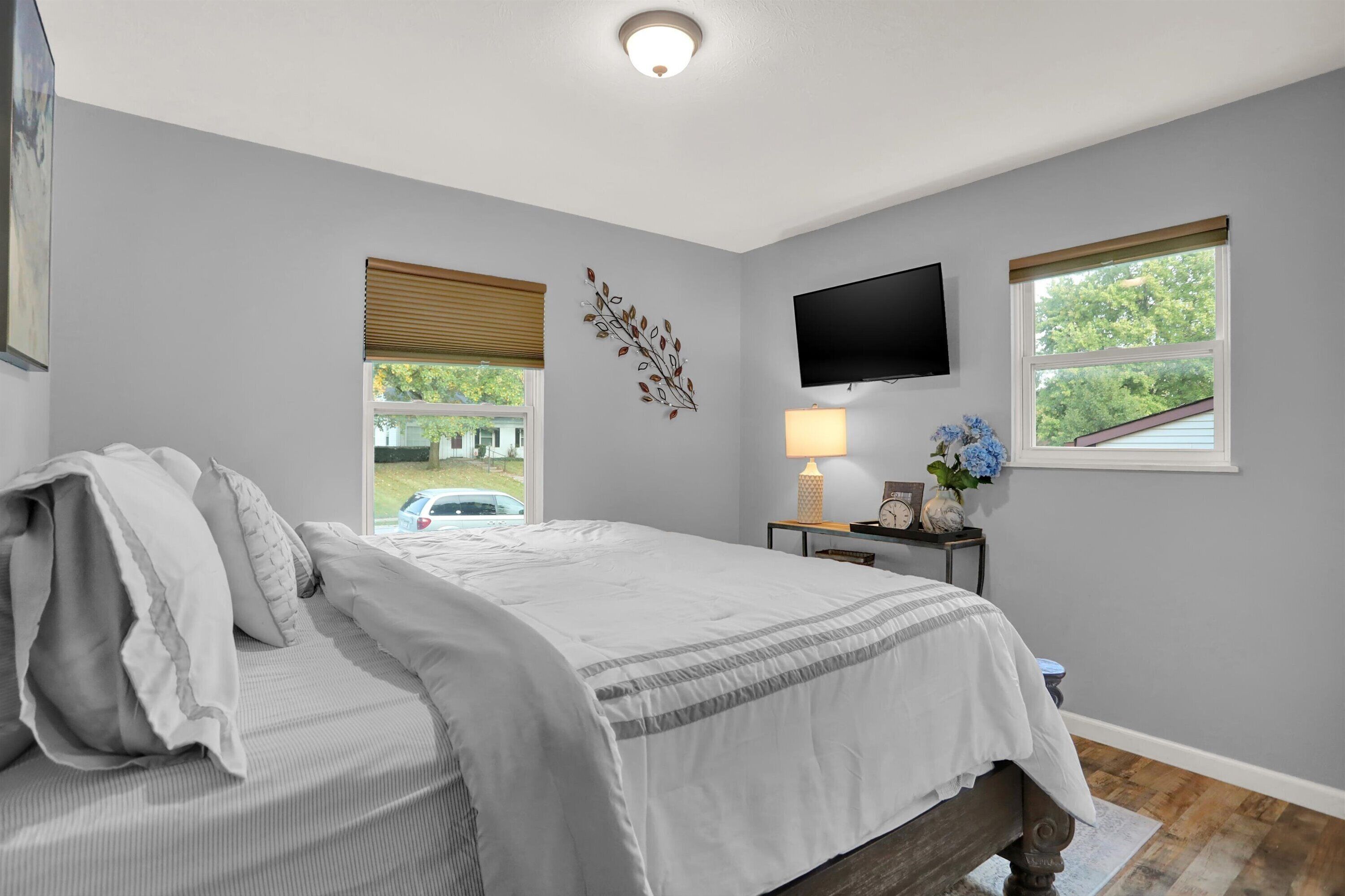 A bedroom with a bed, a TV mounted on the wall, a window with blinds, and a lamp on a bedside table.
