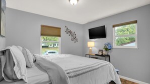 A bedroom with a bed, a TV mounted on the wall, a window with blinds, and a lamp on a bedside table.
