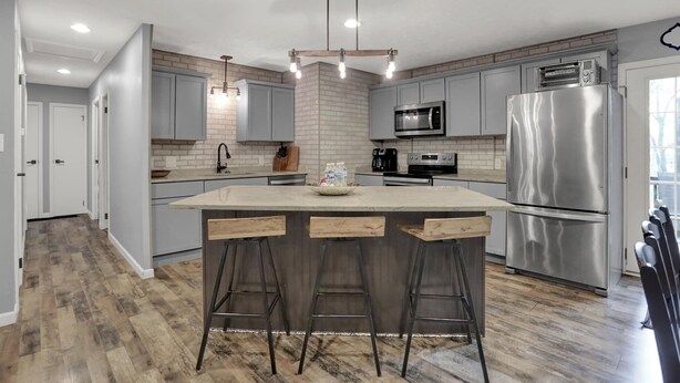 A modern kitchen with grey cabinets, a central island with stools, stainless steel appliances, and a brick accent wall.