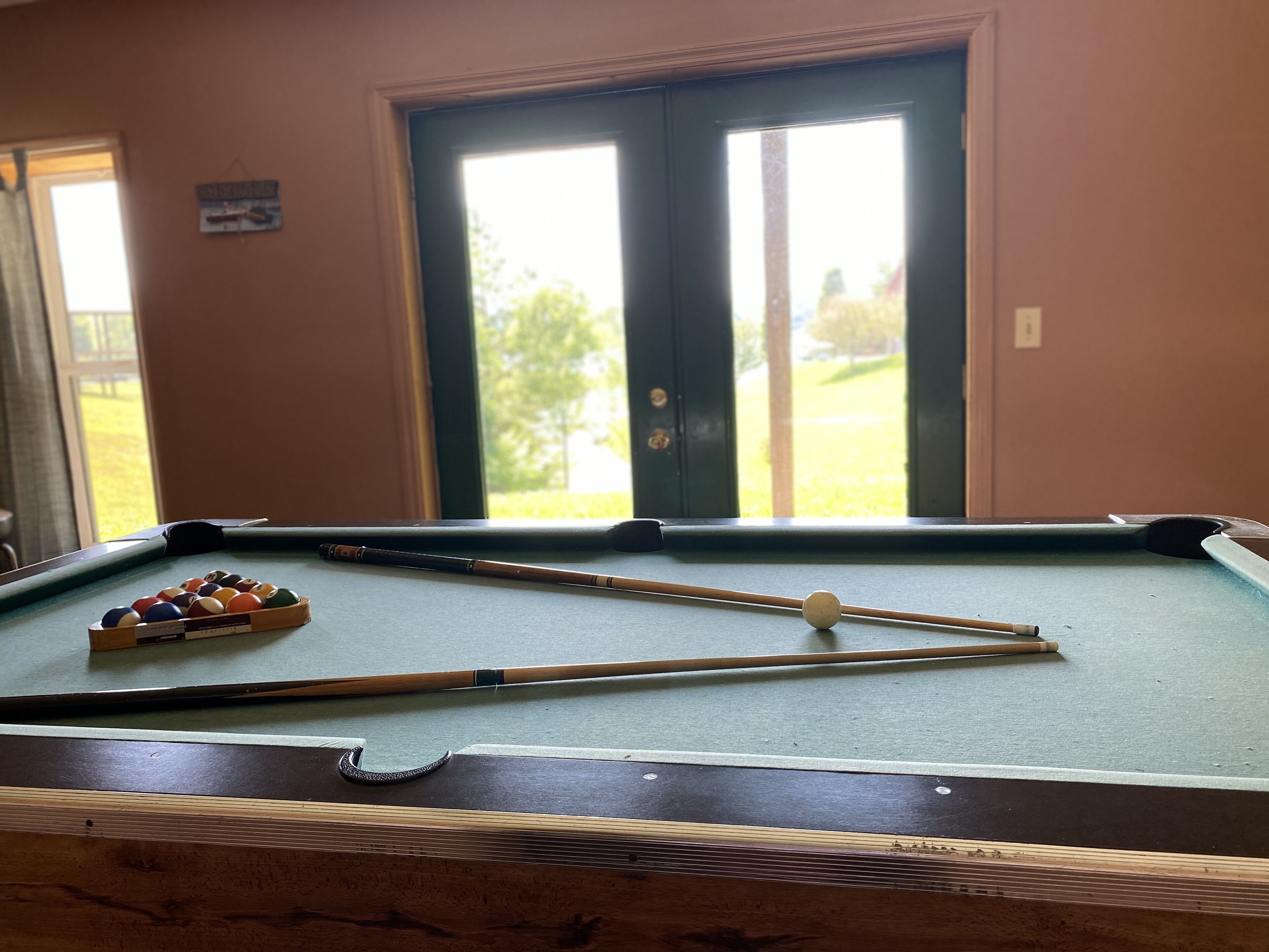 A pool table with pool balls and cues, a window with a view of greenery, and a door with a sign on the wall.