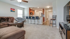 A modern living room with a sofa, a coffee table, and a TV stand. There is a kitchen area with a bar stool and a ceiling fan. - Corporate, Lackland, Sea World, Alamo, Fiesta, Riverwalk BMT graduation (San Antonio)