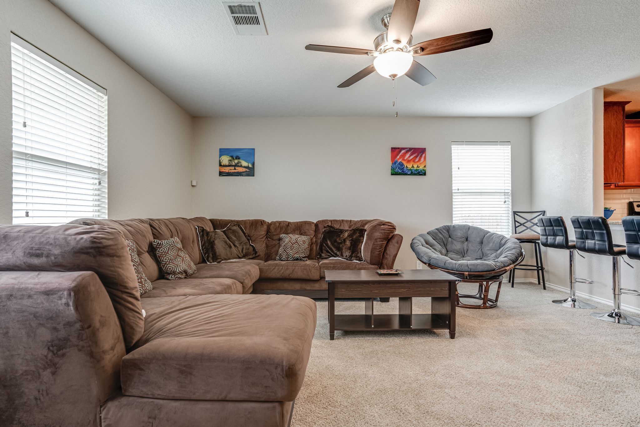 A living room with a sectional sofa, a round chair, a coffee table, and a ceiling fan. There are two windows with blinds, a painting on the wall, and a kitchen area in the background.