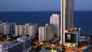 A high-rise hotel with a prominent 'H' logo, surrounded by other buildings and a view of the ocean.