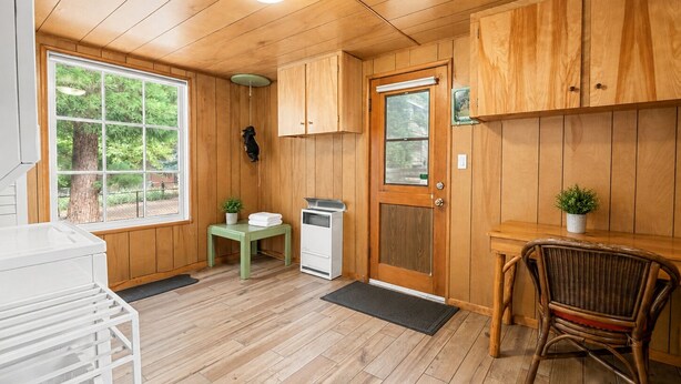 A wooden-paneled room with a window, a door, a small table with a plant, a chair, and a white appliance. - Bear Slope View - Close to Slopes, Pet Friendly, Fenced Yard, Hot Tub (Big Bear Lake)