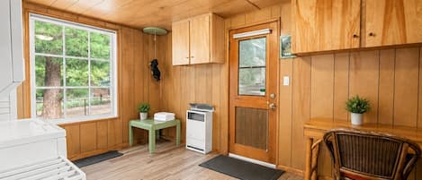 A wooden-paneled room with a window, a door, a small table with a plant, a chair, and a white appliance.