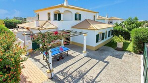 A white building with a tiled roof and green shutters, a courtyard with a ping pong table, and surrounding greenery. - Villa White Sands - Private Oasis, Heated Pool, Beaches & Strip Nearby (Albufeira)