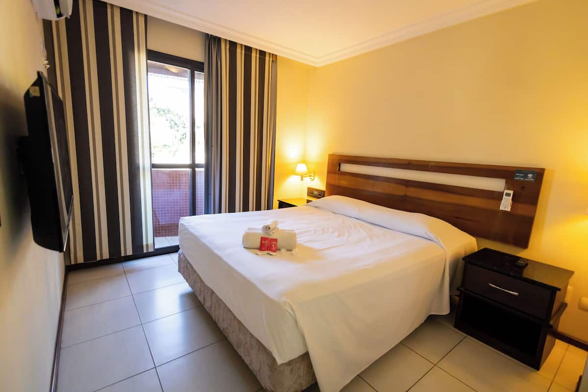 Une chambre d’hôtel avec un grand lit, une table de chevet avec une lampe, un téléviseur fixé au mur et une porte-fenêtre coulissante avec des rideaux.