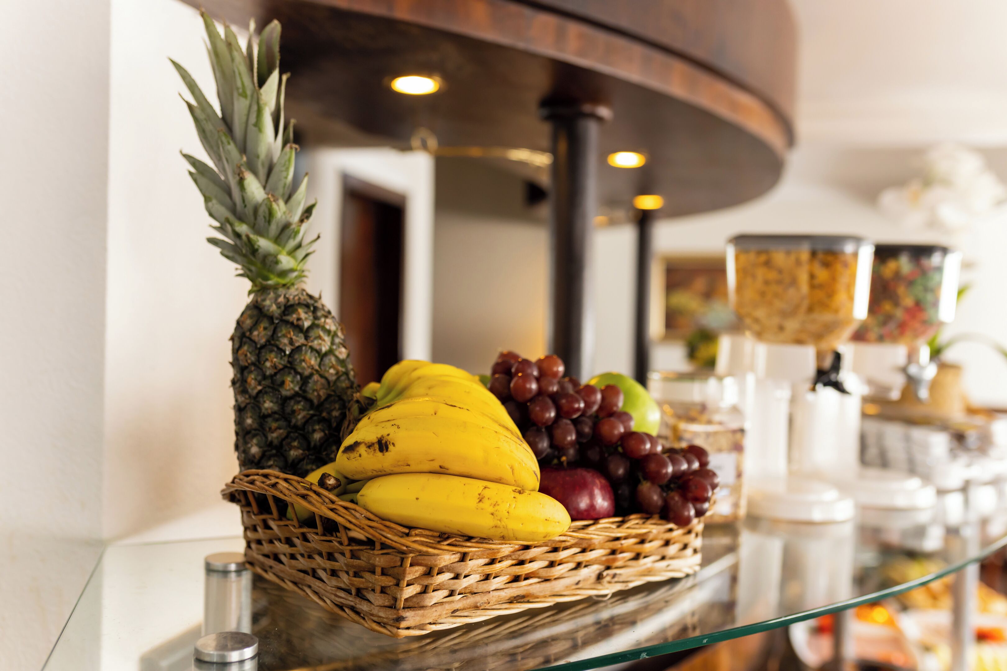 Una cesta de frutas sobre una mesa de vidrio con piña, plátanos, uvas y manzanas.
