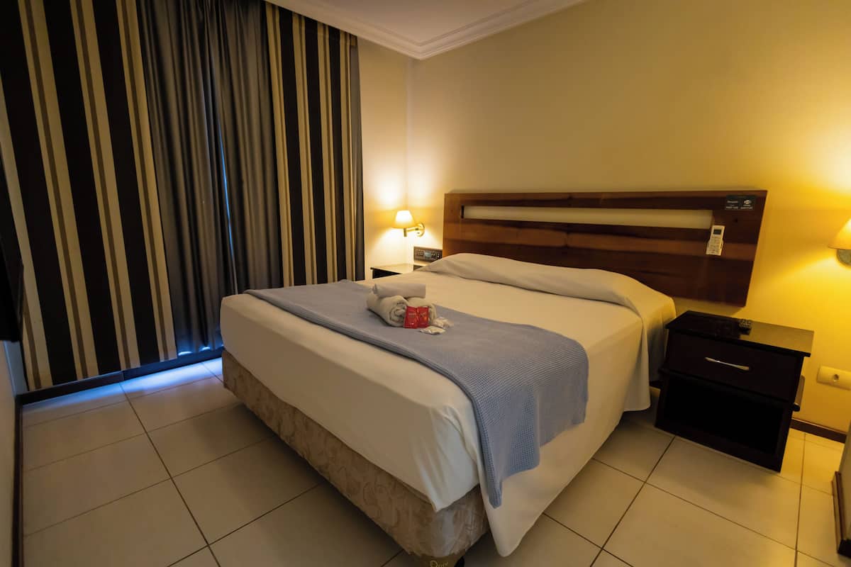 Une chambre d’hôtel avec un grand lit, une table de chevet, une lampe et une fenêtre avec des rideaux.