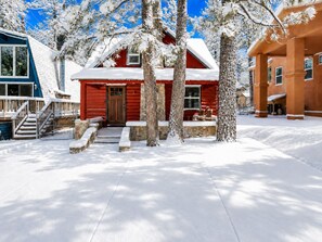 A snow-covered wooden cabin with a stone foundation, a red door, and a porch with outdoor seating.
