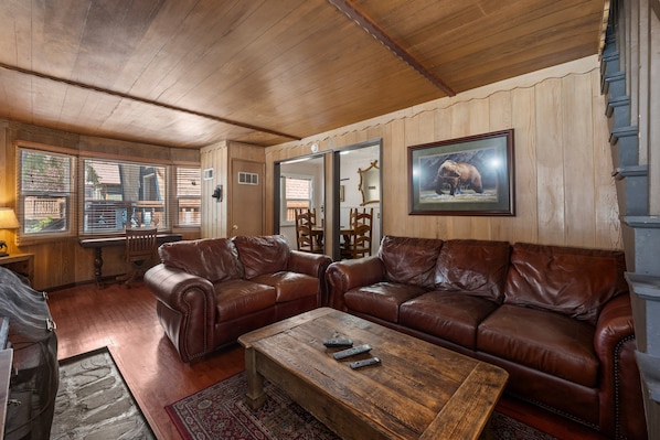 A cozy living room with leather sofas, a wooden coffee table, and a large window with blinds.
