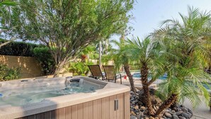Ett bubbelbad omgivet av träd och en pool, med solstolar och en stenmur i bakgrunden.