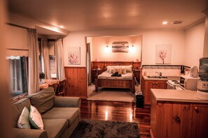 Cozy room with a sofa, bed, and kitchenette, featuring a wooden table, lamp, and artwork.