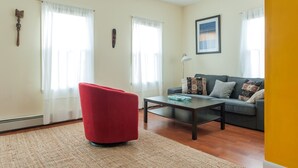 A living room with a red chair, a grey sofa, and a coffee table. There are two windows with white curtains, a lamp, and a framed picture on the wall.