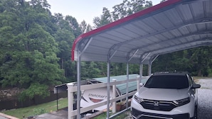 A covered carport with a white car parked inside, surrounded by trees and a gravel driveway.