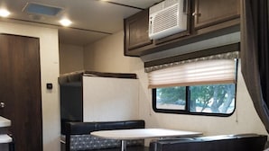 A compact RV interior with a bed, kitchenette, and a window with a view.