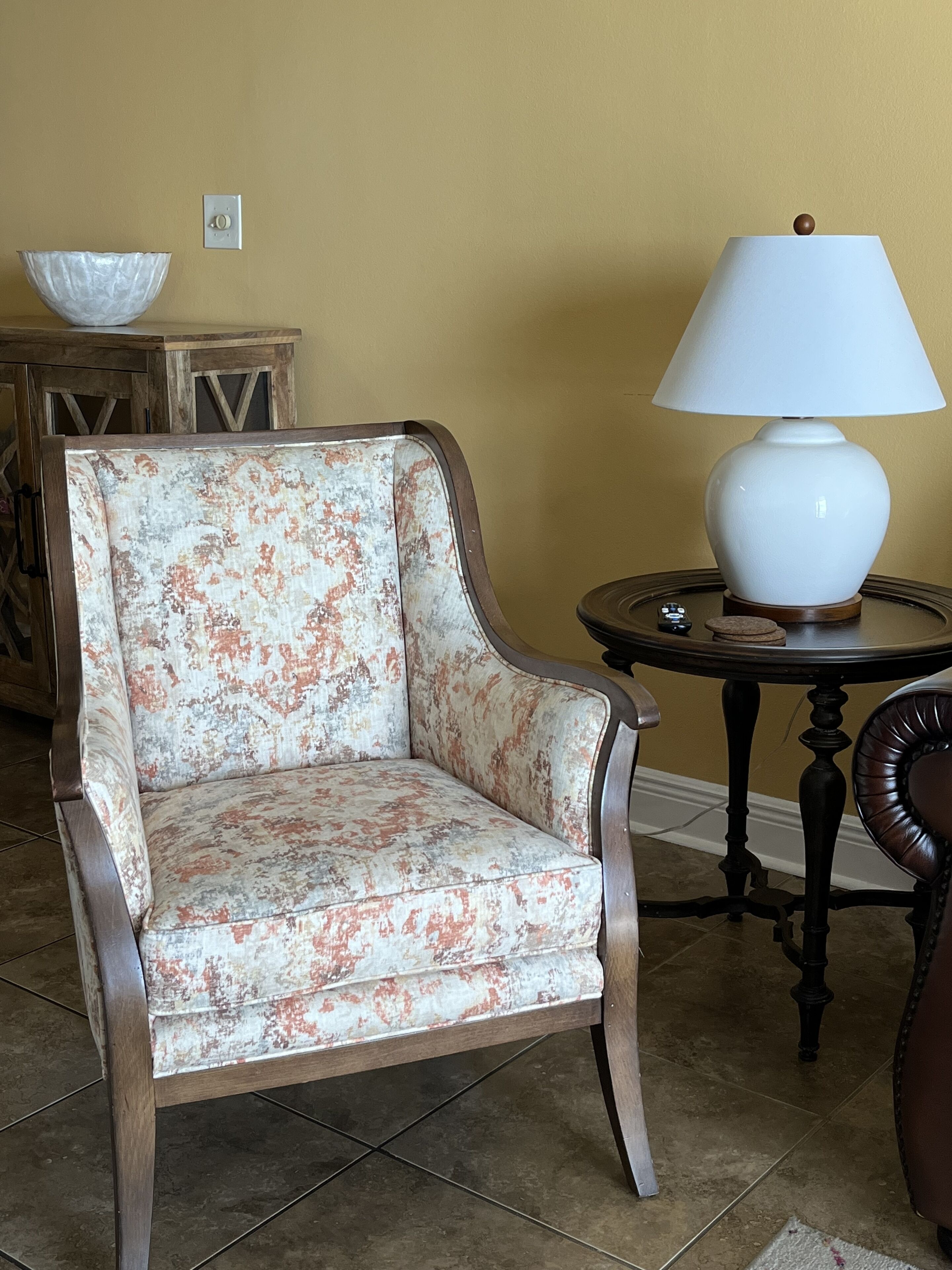 A chair with a floral pattern, a table with a lamp, and a bowl on a wooden cabinet.